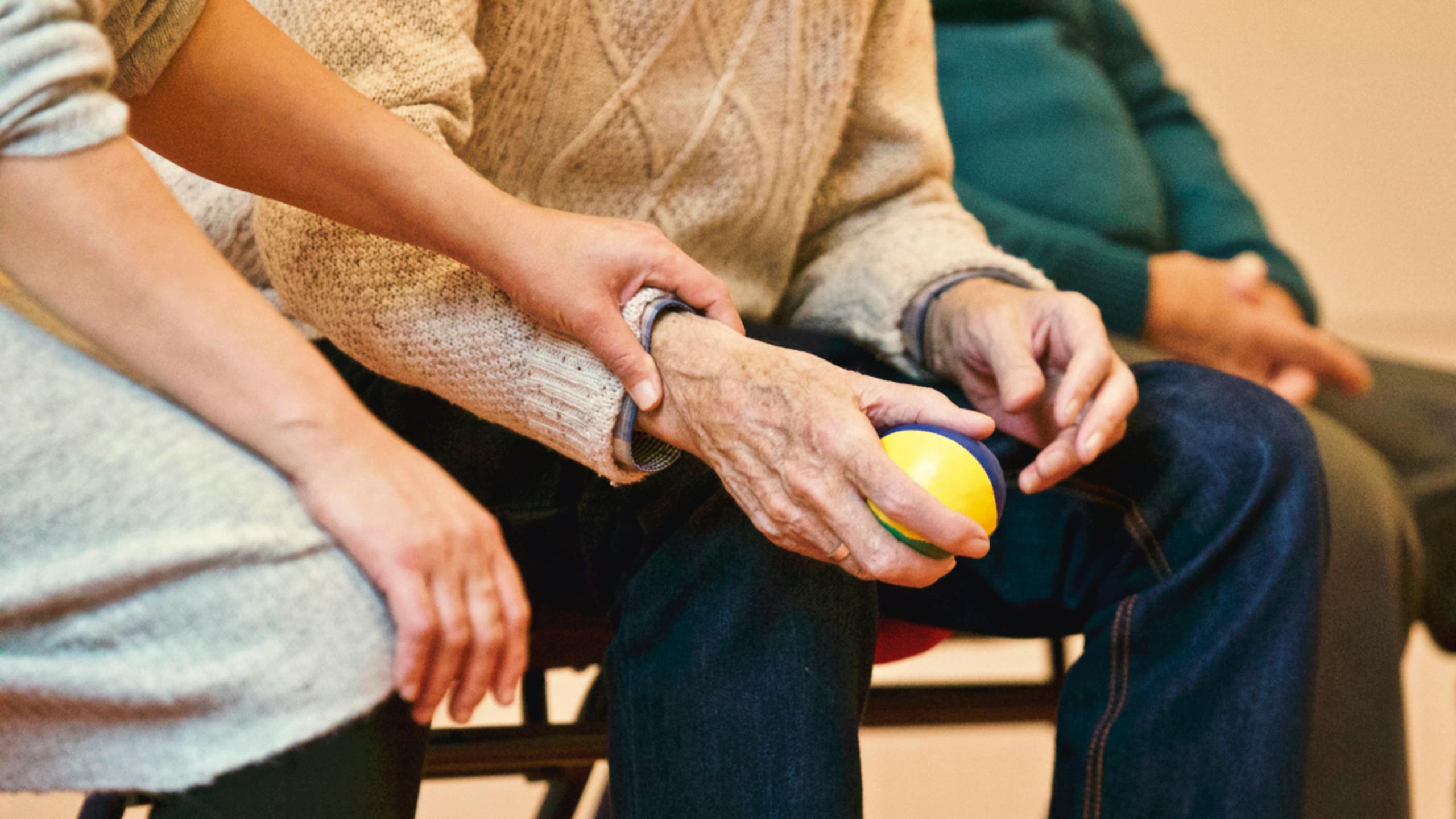 Manos de un cuidador ayudando a un anciano a agarrar una pelota durante una práctica de rehabilitación